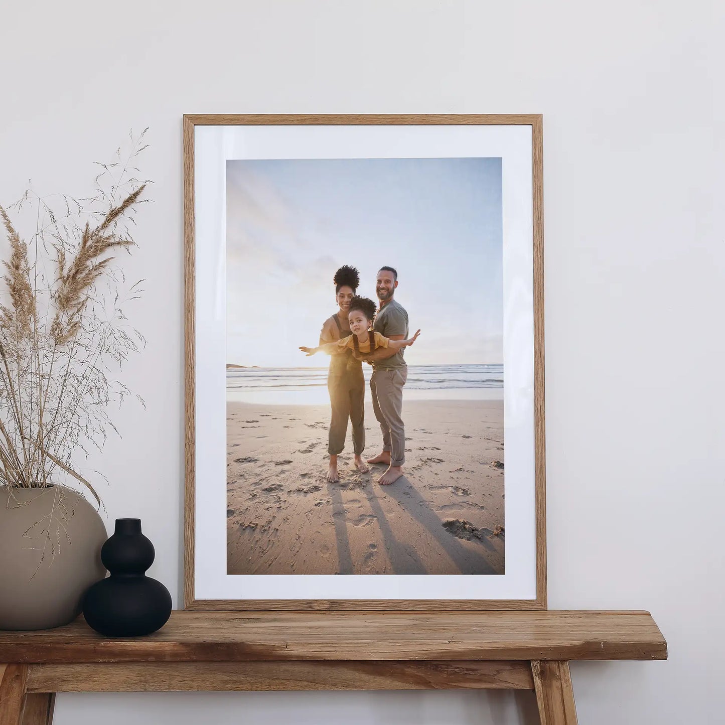 Light wood framed large-format vertical photo print of a family standing on a sandy beach at sunset, displayed on a wooden bench beside decorative vases.