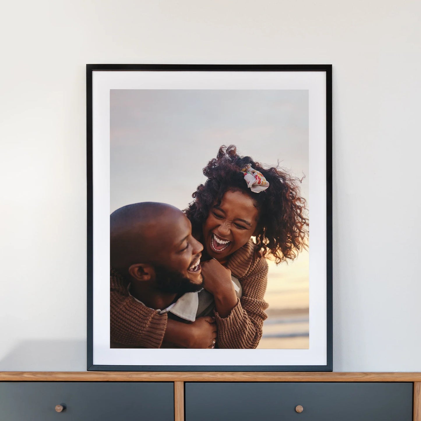 Black framed large-format vertical photo print of a couple laughing together on a beach, displayed on a mid-century style cabinet against a neutral wall.