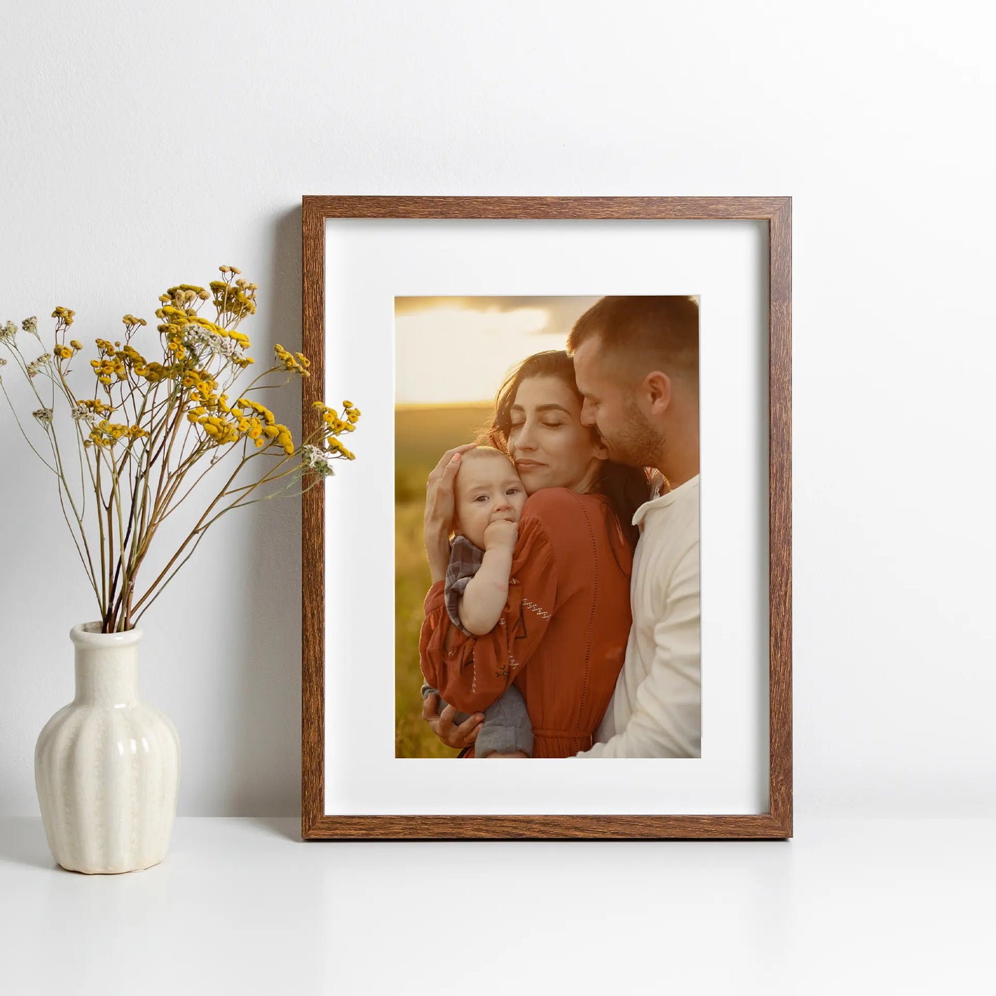 Wood framed large-format vertical photo print of parents holding a baby during golden hour outdoors, displayed next to a ceramic vase with yellow flowers.