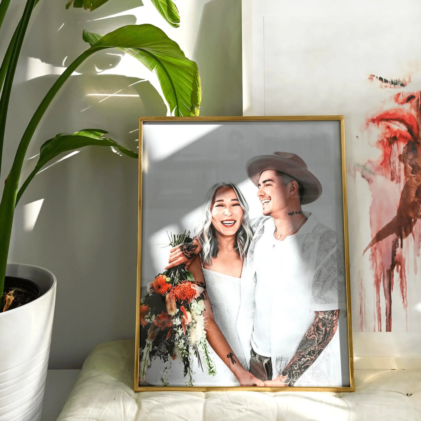 Gold framed large-format vertical photo print of a smiling couple holding a bouquet, displayed near a large green plant with soft window light.