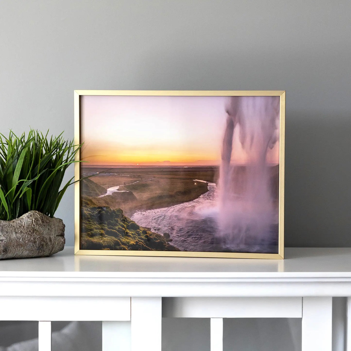 Gold framed large-format horizontal landscape print of a waterfall cascading beside a river at sunset, displayed on a white console table next to a green plant.