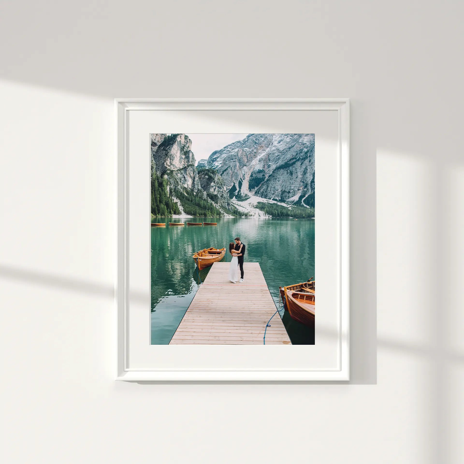 Large format photo print in a frame, showing a couple embracing on a wooden dock over a mountain lake, surrounded by alpine scenery and boats.