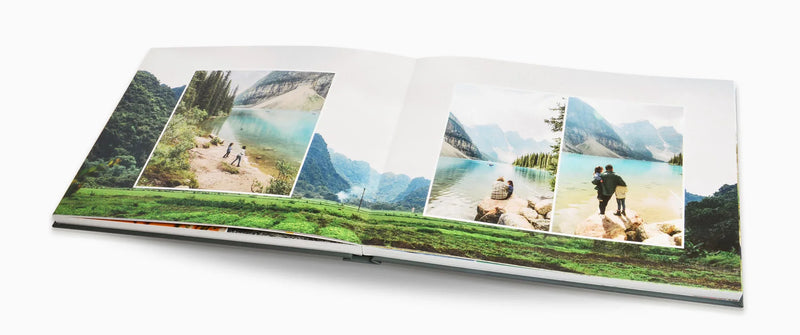 Open layflat photo book displaying travel photos of people by a turquoise mountain lake and scenic landscape spread across both pages with full-bleed background image.