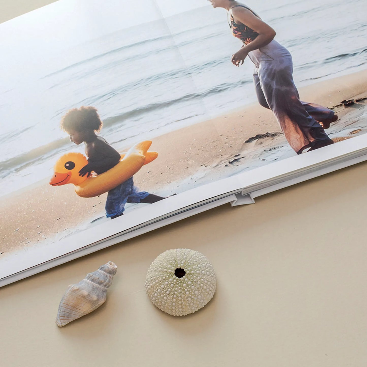 Layflat photo book open to full-bleed beach image of child with inflatable float and adult walking along shoreline, placed beside seashells.