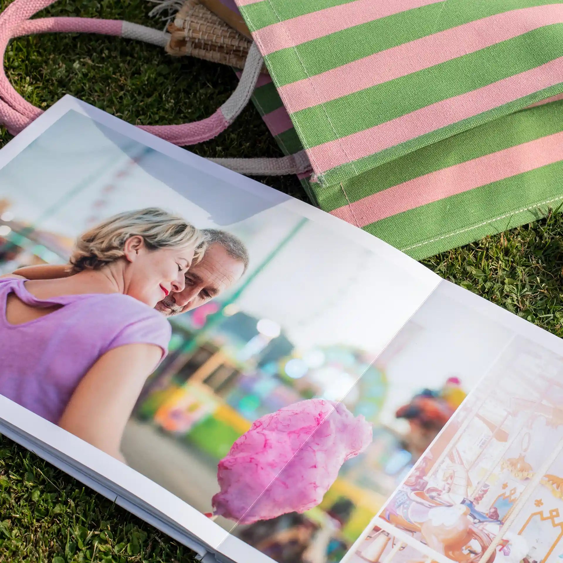 Layflat photo book open to full-bleed image of couple at outdoor fair holding cotton candy, displayed on grass beside striped tote bag.