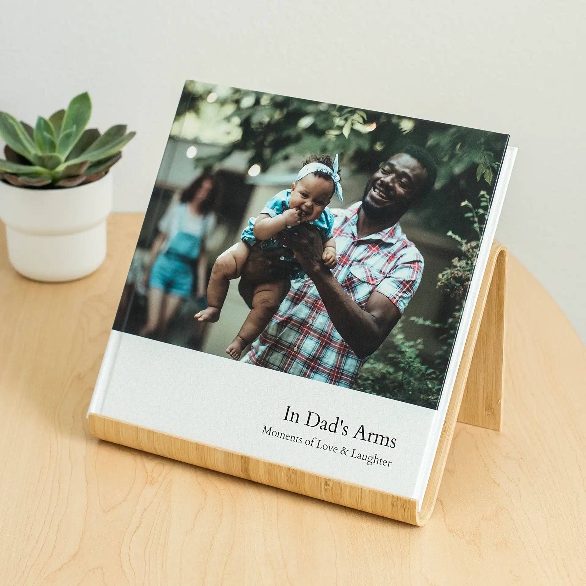 Layflat photo book titled 'In Dad's Arms: Moments of Love & Laughter' displayed upright on a wooden stand on a table beside a small succulent, featuring a cover photo of a father laughing as he lifts his smiling baby outdoors