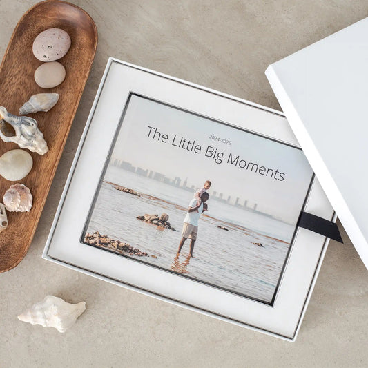 Layflat photo book inside presentation box featuring cover image of adult carrying child at beach, with printed title text and coastal background.