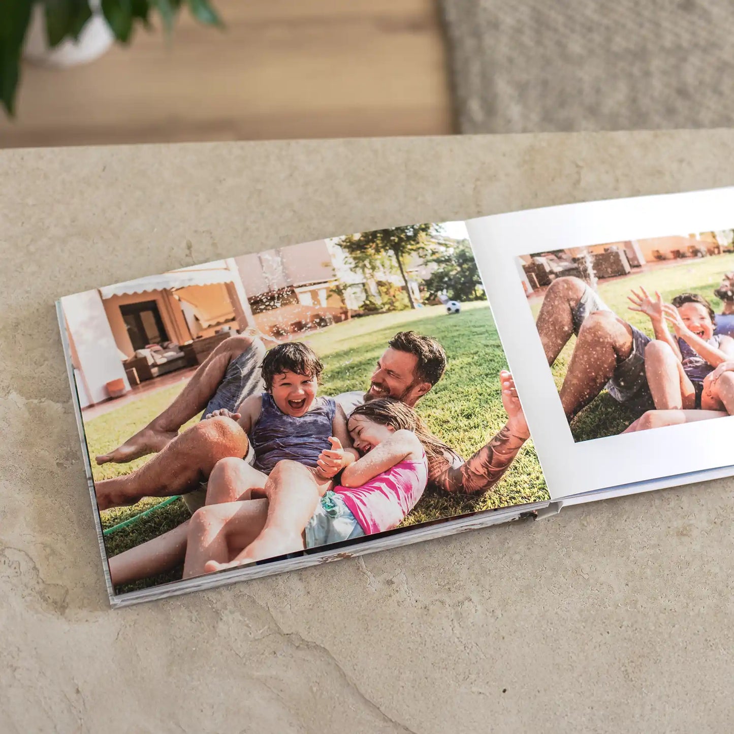 Open layflat photo book on a stone surface showing a seamless spread of a father and his two young children laughing together while playing in backyard sprinklers on a sunny summer day
