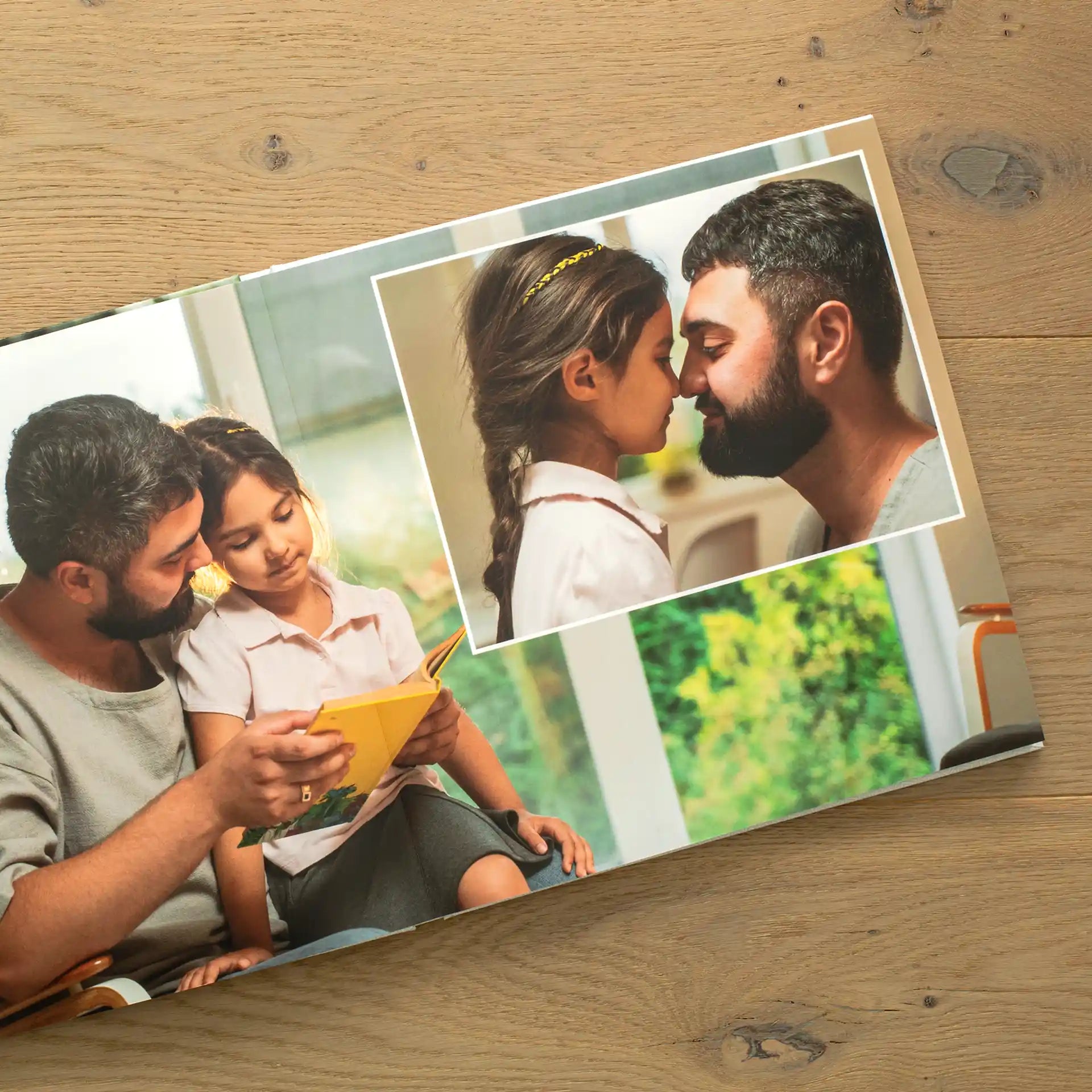 Open layflat photo book on a wooden surface displaying a tender spread of a father reading with his young daughter, alongside a close-up photo of the pair touching foreheads affectionately