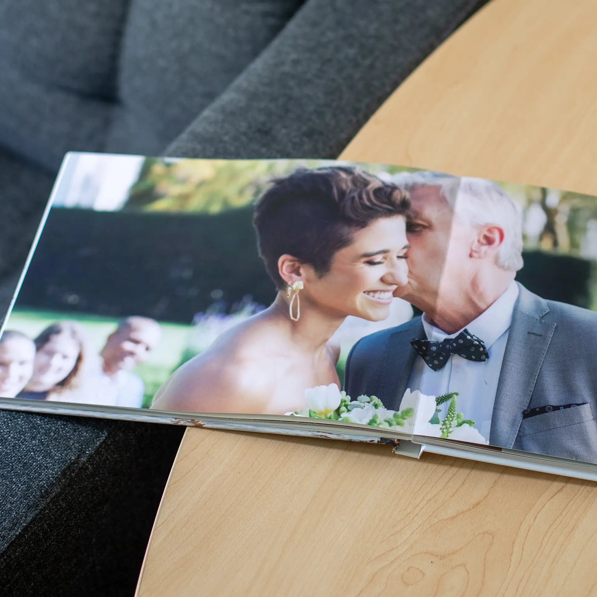 Open layflat photo book resting on a wooden coffee table showing a full-bleed wedding photo of a father of the bride kissing his daughter on the cheek as she smiles holding her bouquet
