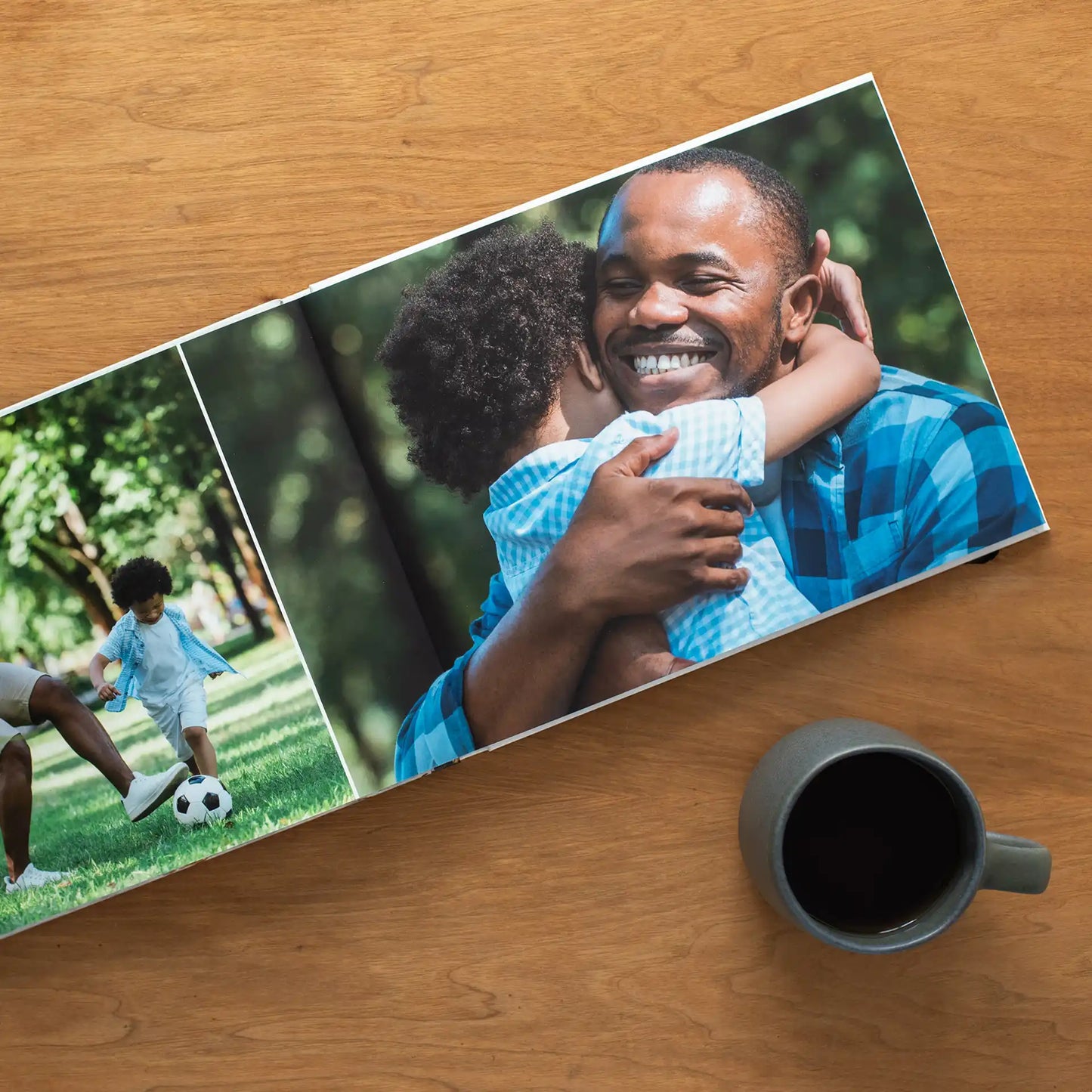 Open layflat photo book on a wooden desk beside a coffee mug, displaying a spread of a father and young son playing soccer in a park alongside a full-page photo of the pair sharing a warm embrace
