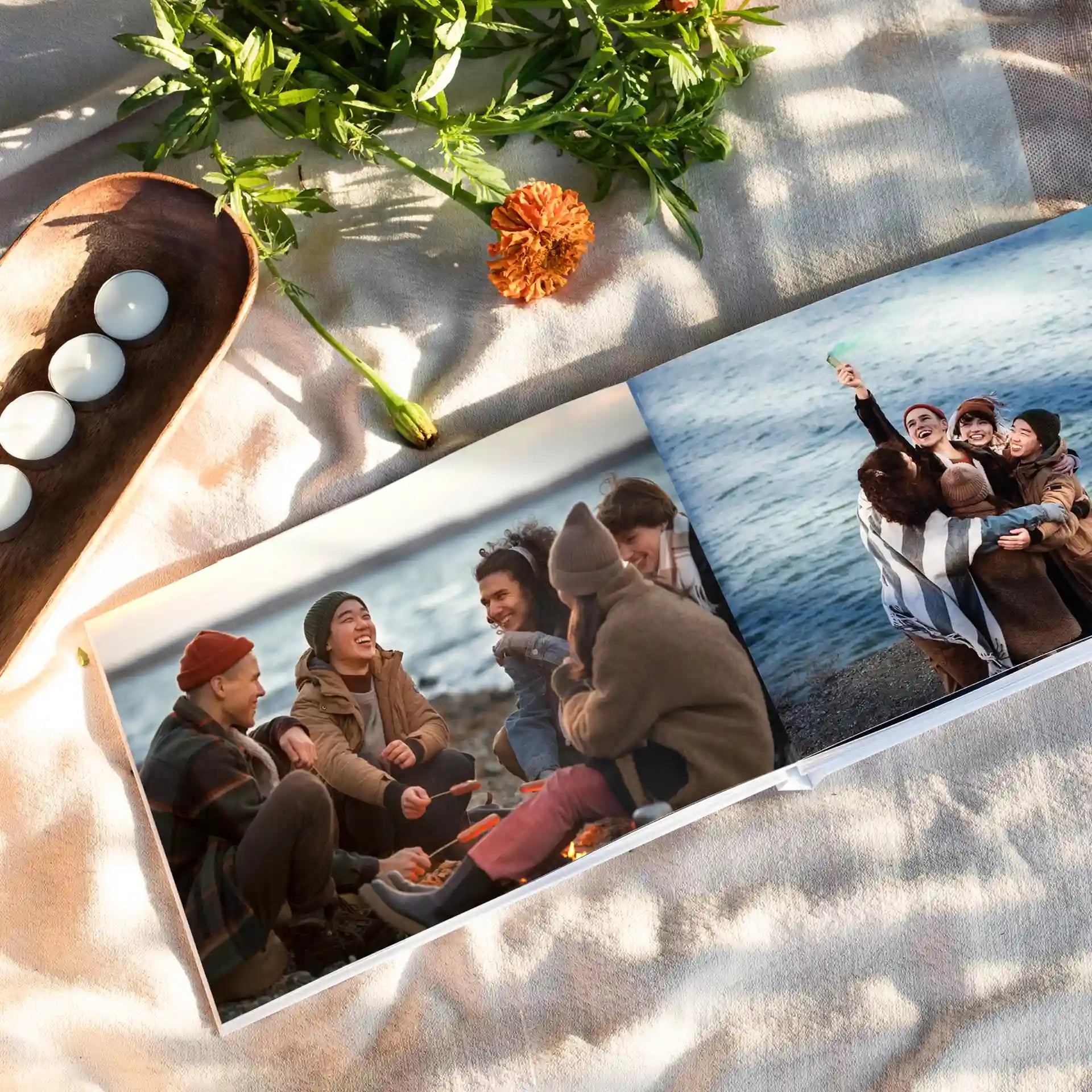 Open layflat photo book lying on a light textured blanket, showing two spread photos of friends on a beach trip — one group gathered around a fire, another cheering by the ocean — surrounded by candles and greenery.