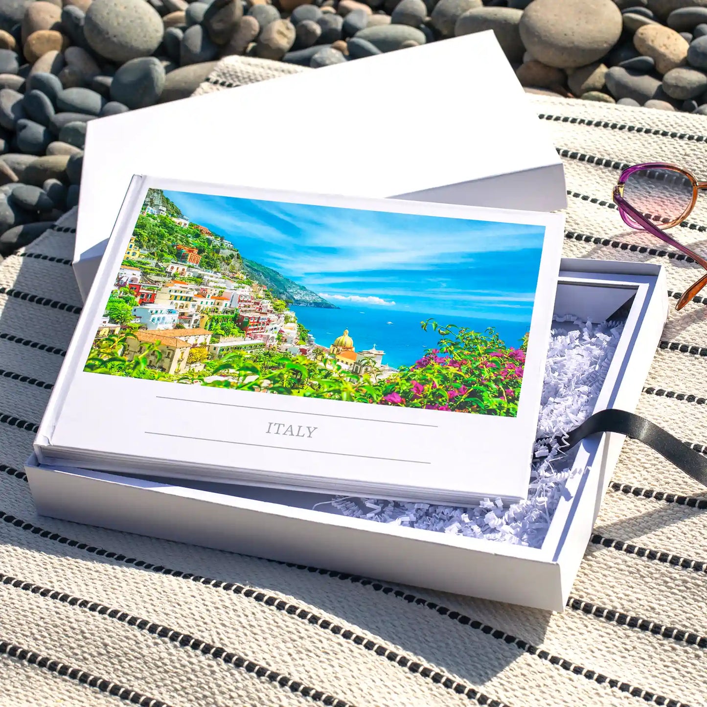 Layflat photo book with travel image of coastal Italy on cover, placed inside open gift box with tissue paper on outdoor picnic blanket.