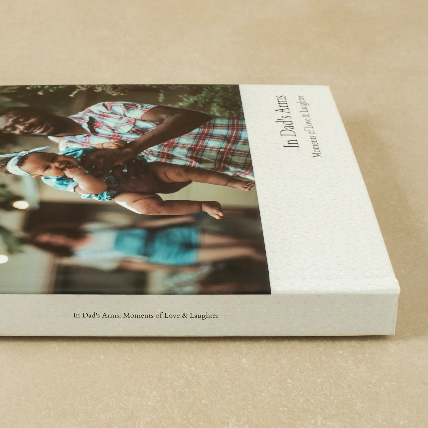 Close-up detail shot of a layflat photo book spine and cover showing the printed title 'In Dad's Arms: Moments of Love & Laughter' alongside a photo of a father lifting his baby in the air