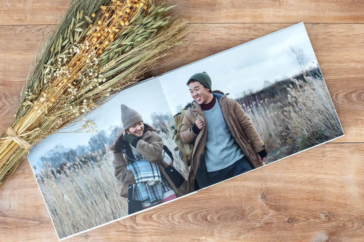 An open layflat photo book with a seamless two-page photo of a smiling couple in a field, beside a bundle of dried grasses.