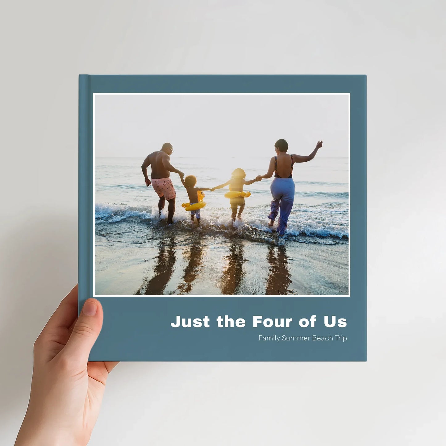 10x10 square hardcover photo book showing a teal cover with a family of four entering the ocean, photographed from behind.