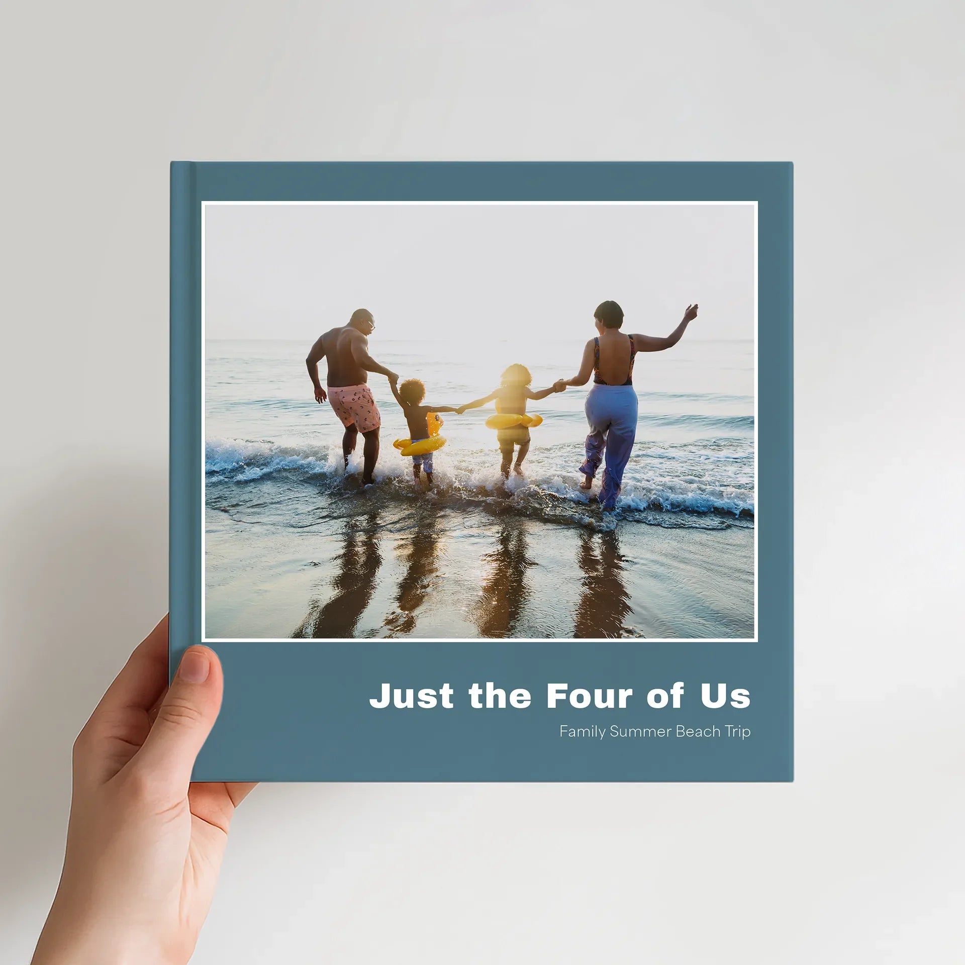 10x10 square hardcover photo book showing a teal cover with a family of four entering the ocean, photographed from behind.