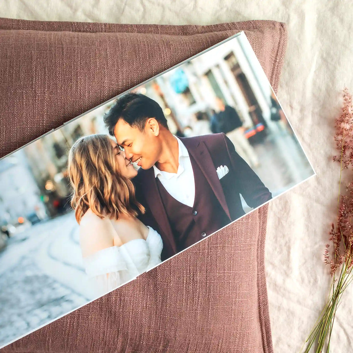 Photo book with a wedding photo of a couple touching noses on a city street, lying on a mauve linen pillow beside dried grasses.
