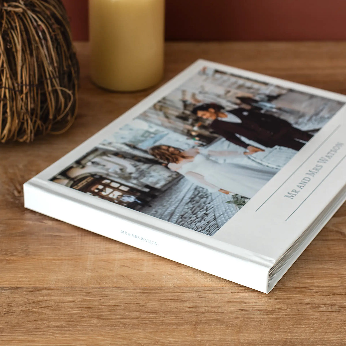 Closed layflat photo book with a custom wedding cover reading 'Mr and Mrs Watson,' displayed on a wood table with candles.