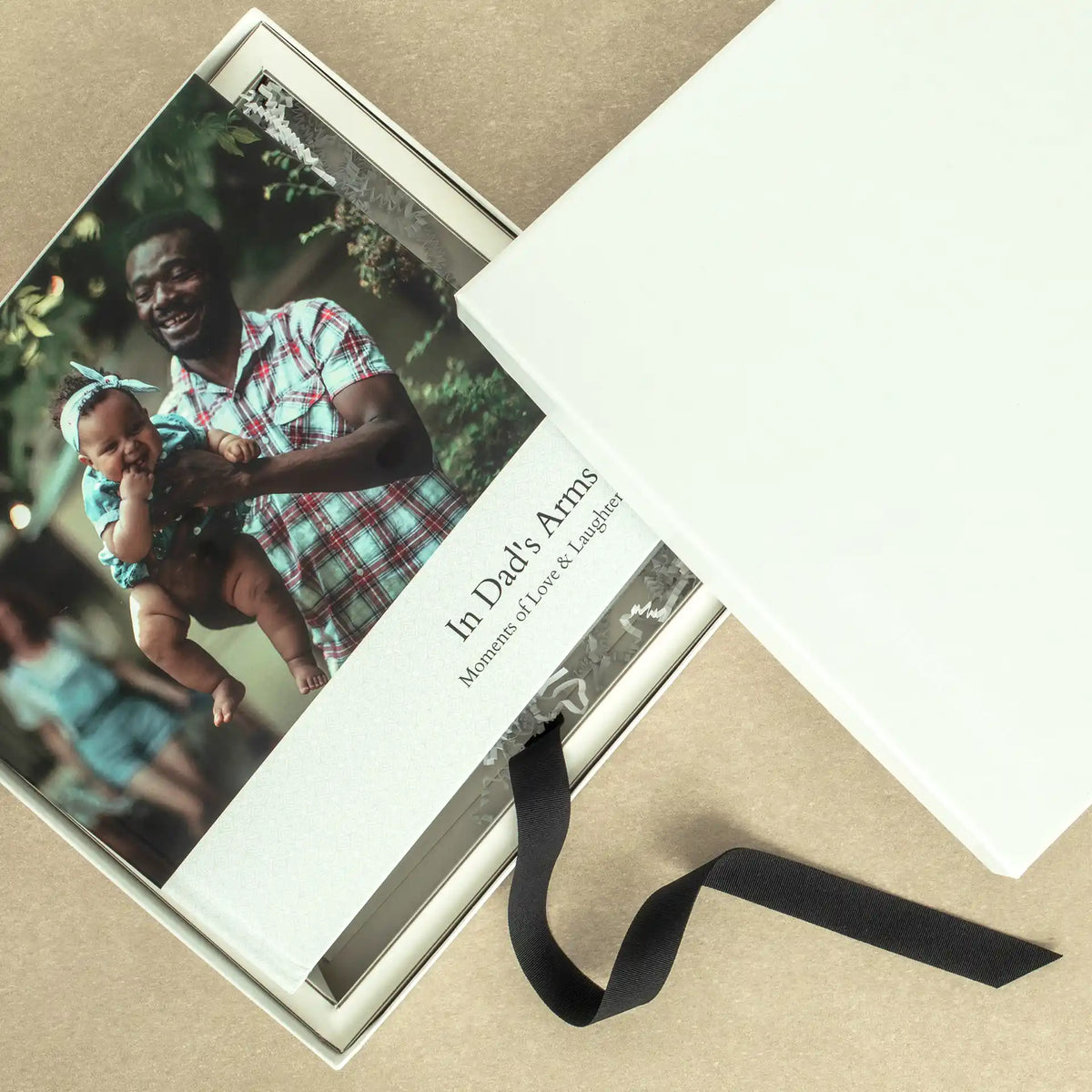 Layflat photo book titled 'In Dad's Arms: Moments of Love & Laughter,' shown inside a white gift box with black ribbon.