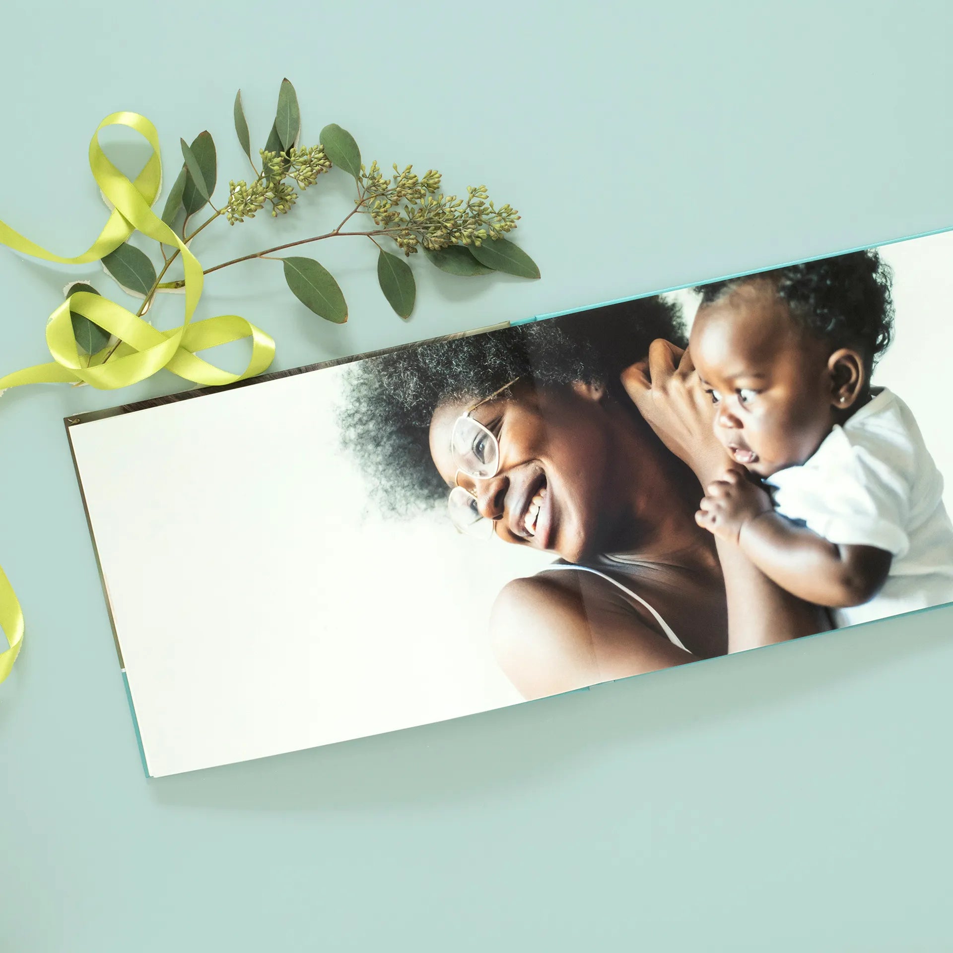 Layflat photo book open to seamless panoramic spread of a person holding a baby, displayed on a soft colored background with ribbon and greenery.