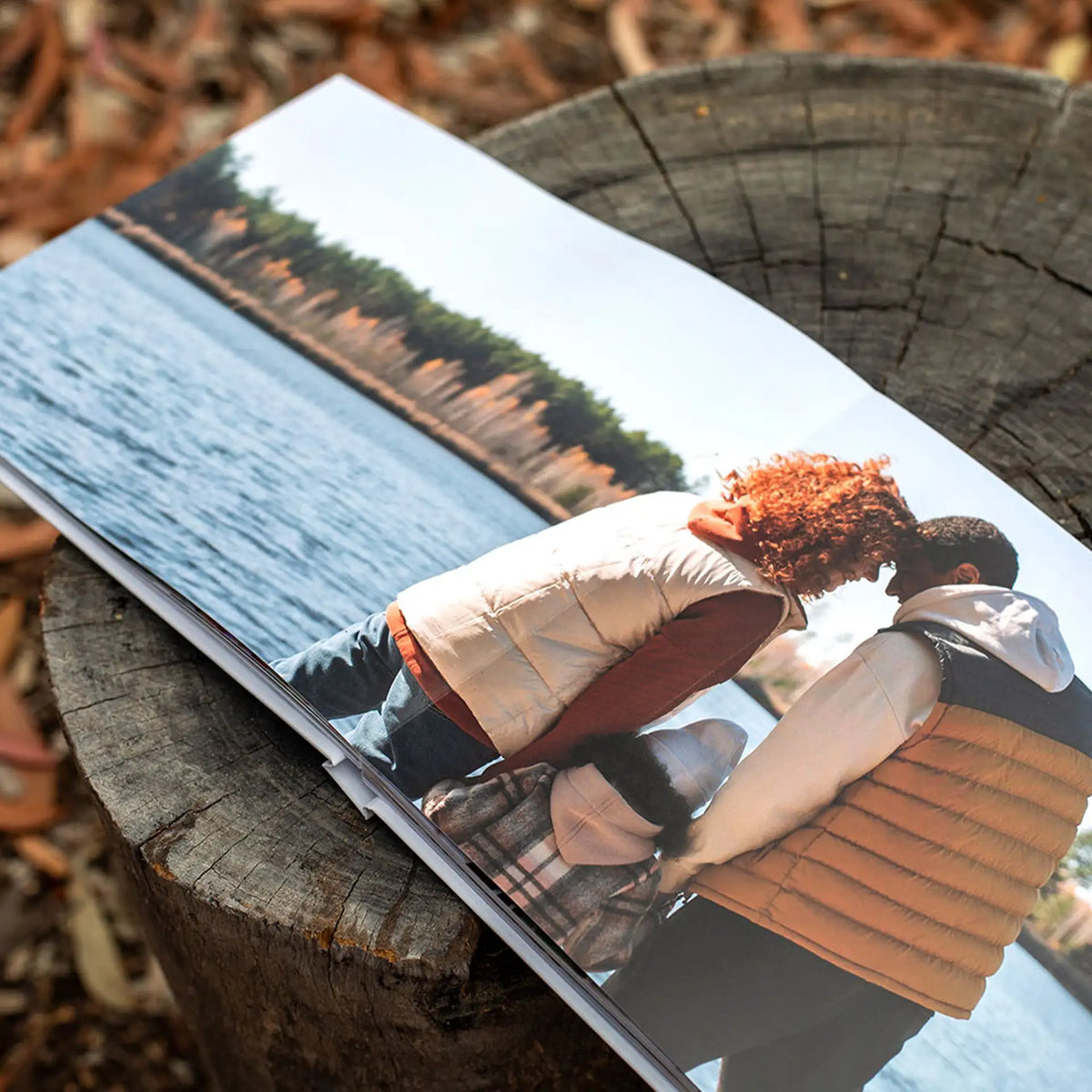 Layflat photo book open on a tree stump outdoors, showing a full-spread photo of a family embracing beside a lake at sunset.