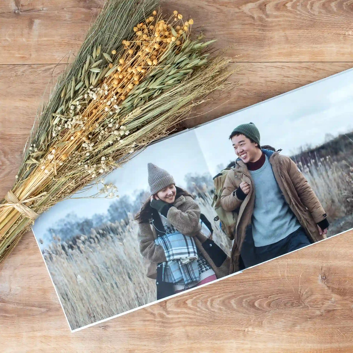 Layflat photo book open to a full-bleed panoramic photo of a couple walking through a golden field, styled next to a dried wheat bouquet.