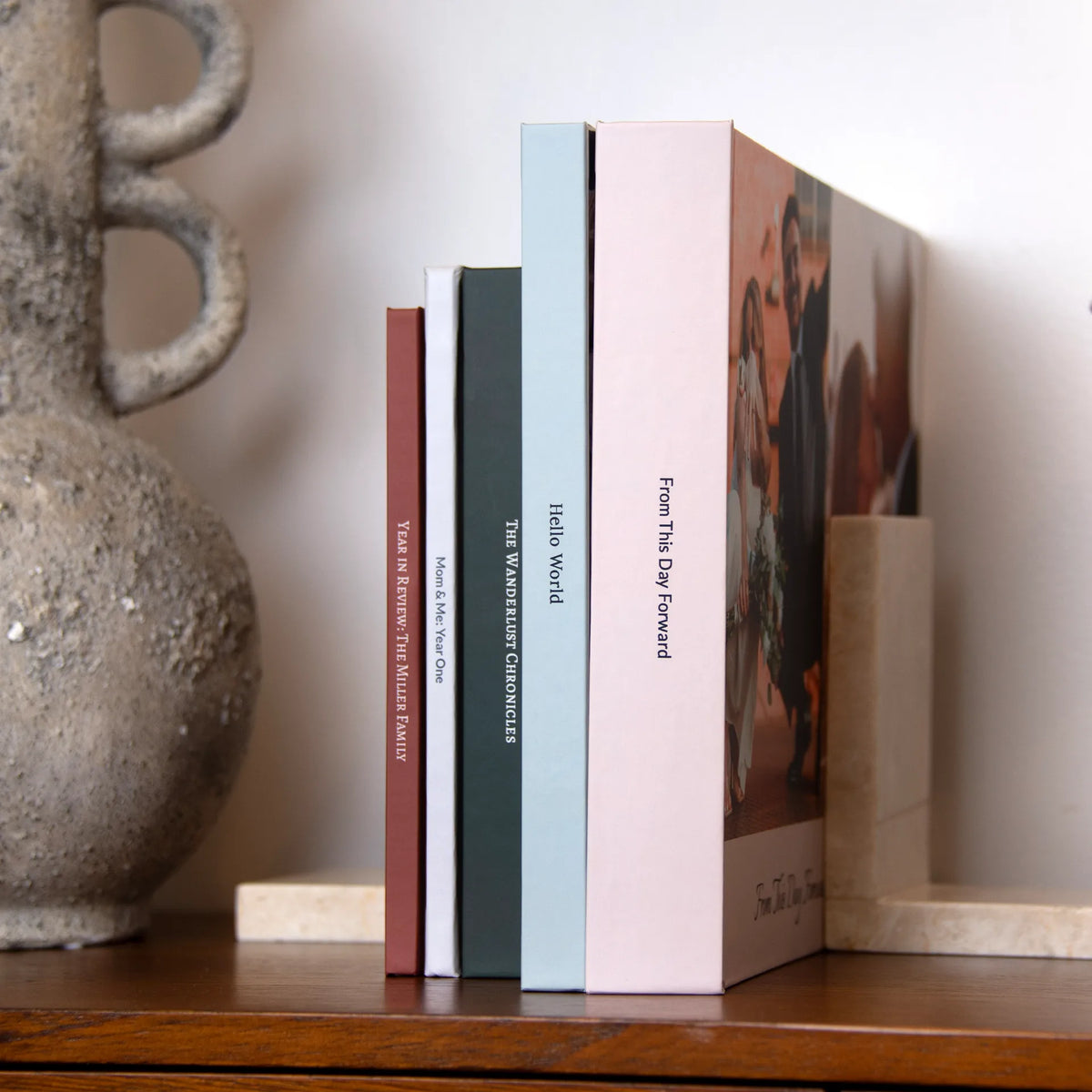 Five layflat photo books in terracotta, white, green, light blue and blush stand on a shelf between marble bookends.