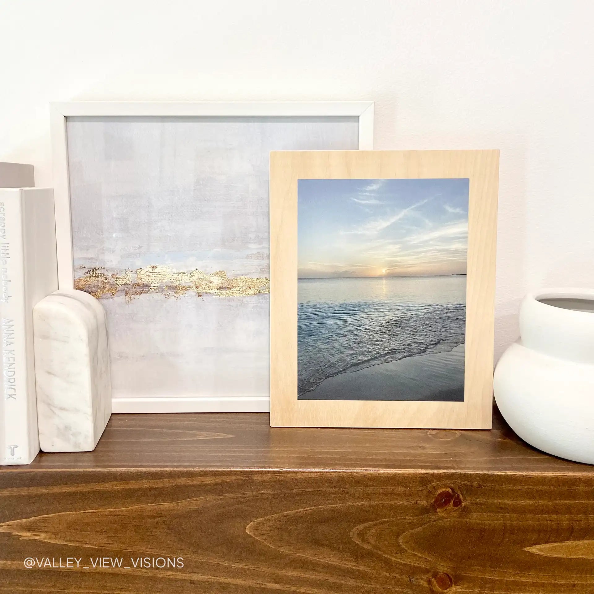Maple wood print of a calm beach shoreline at sunset with soft clouds and gentle waves, displayed on a wooden shelf beside books and vases.
