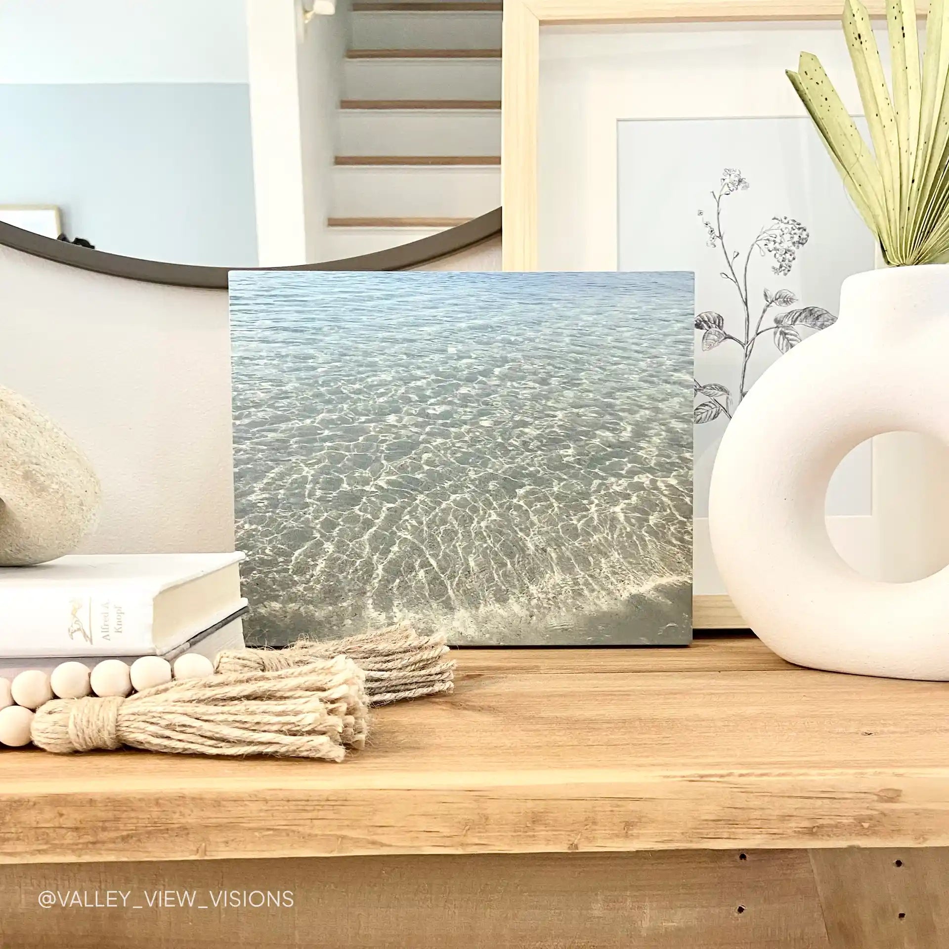 Maple wood print showing clear ocean water with sunlight ripples, displayed on a wooden console table with books and decorative ceramics.