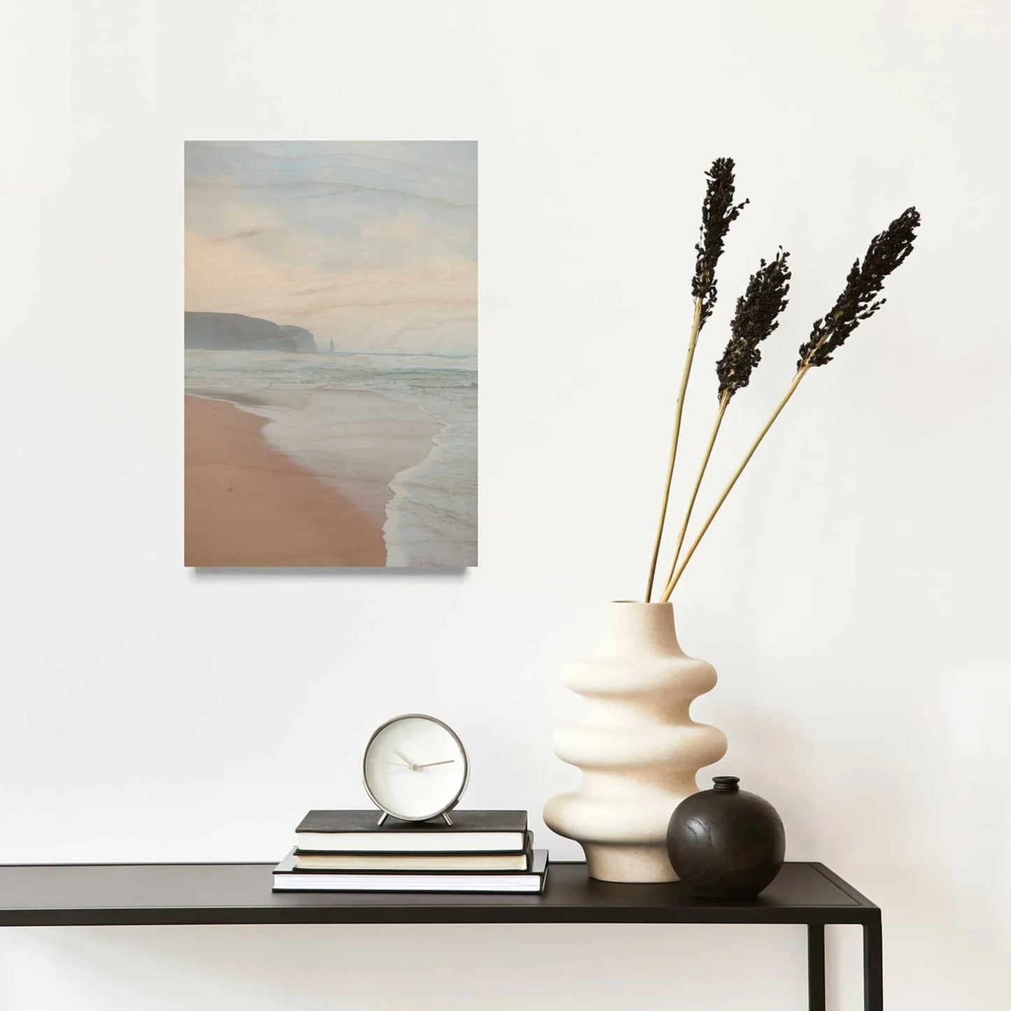 Maple wood print of a sandy beach with coastal cliffs and ocean waves, mounted on a white wall above a console table with decor.