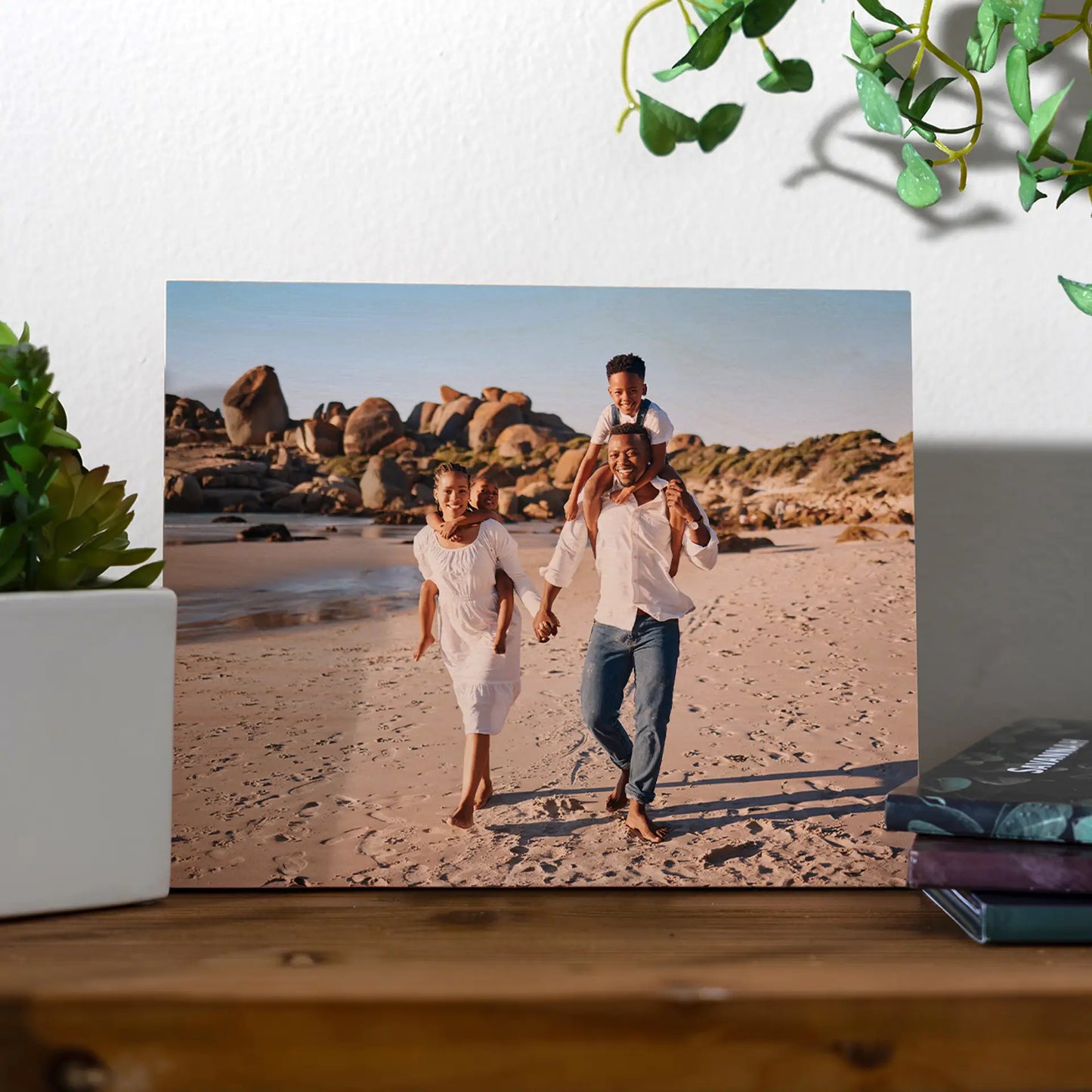 Maple wood print of a family walking barefoot on a sandy beach with rocky shoreline, displayed on a wooden shelf with plants and books.
