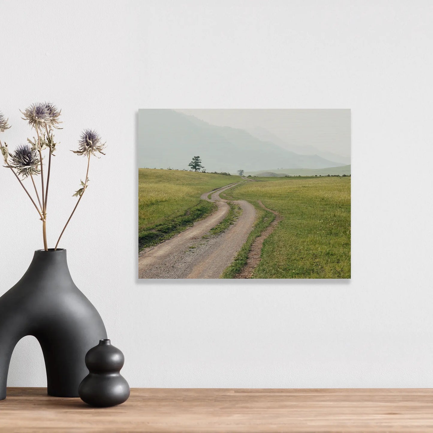 Maple wood print with visible wood texture showing winding dirt path through green fields with distant hills, horizontal orientation, styled above tabletop with ceramic decor.