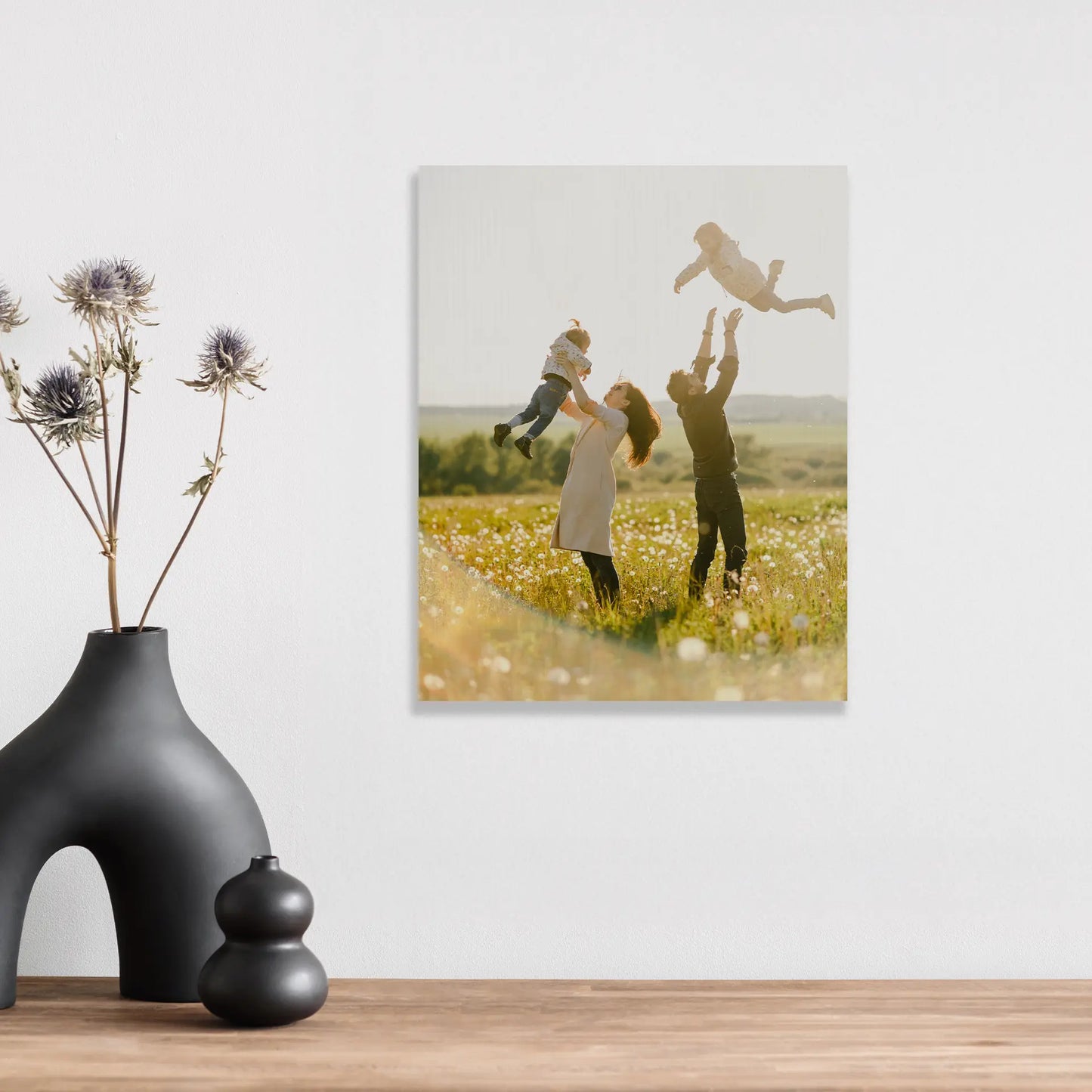 Maple wood print with soft wood texture showing family lifting children in sunlit field, square orientation, displayed above wooden surface with decorative objects.