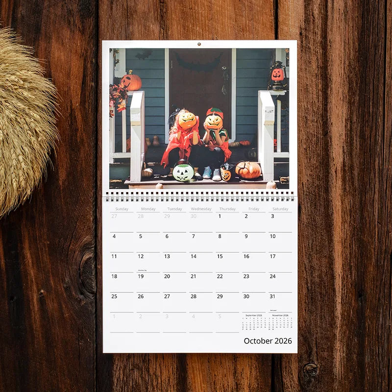 Custom photo wall calendar open to October 2026, showing two kids in pumpkin masks sitting on a decorated front porch, hung on a rustic wood wall.