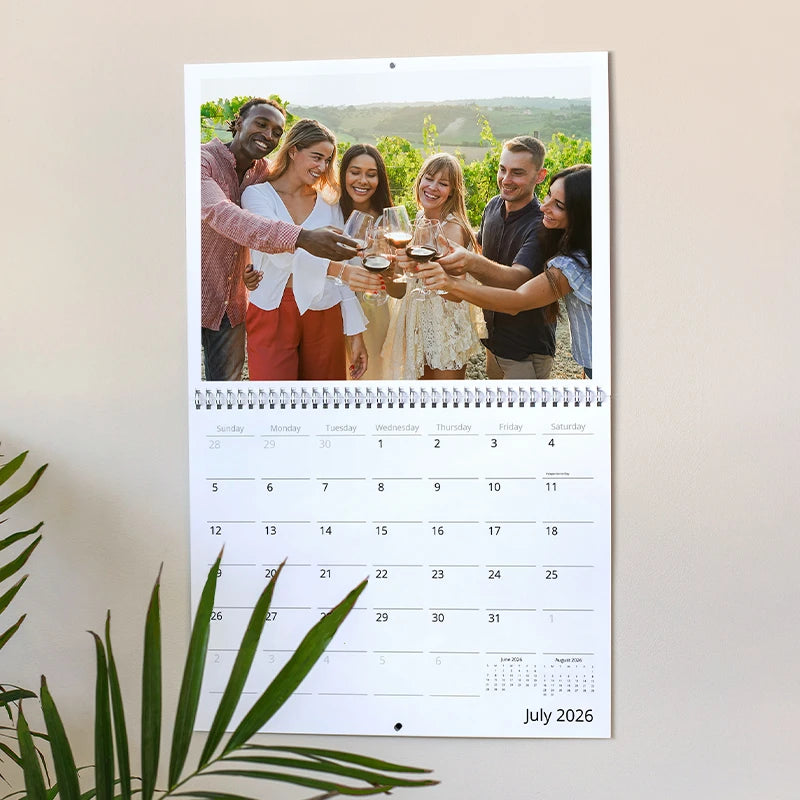 Custom wall calendar showing July 2026 hanging on a wall beside a houseplant, featuring a photo of a group of friends toasting with wine glasses together at a vineyard
