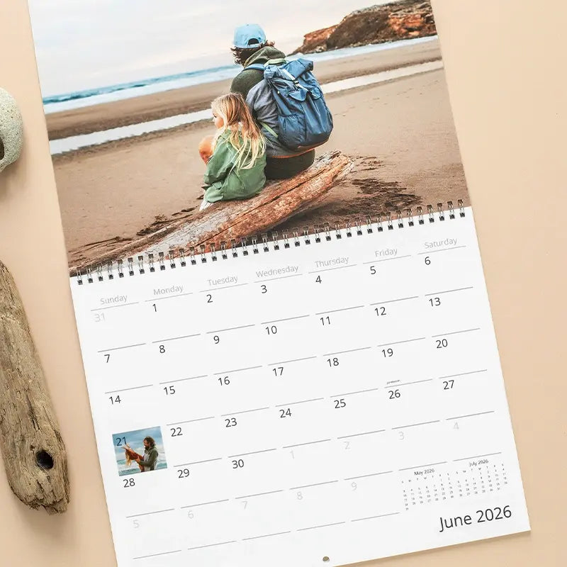 Custom wall calendar showing June 2026 on a beige surface beside driftwood, featuring a photo of a father and young daughter sitting together on a log watching the ocean