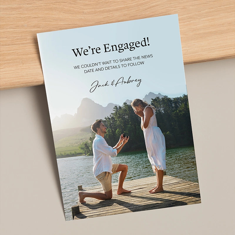 Custom engagement announcement flat card leaning against a wooden surface, reading 'We're Engaged! Jack & Aubrey' with a photo of a man proposing on a dock by a mountain lake