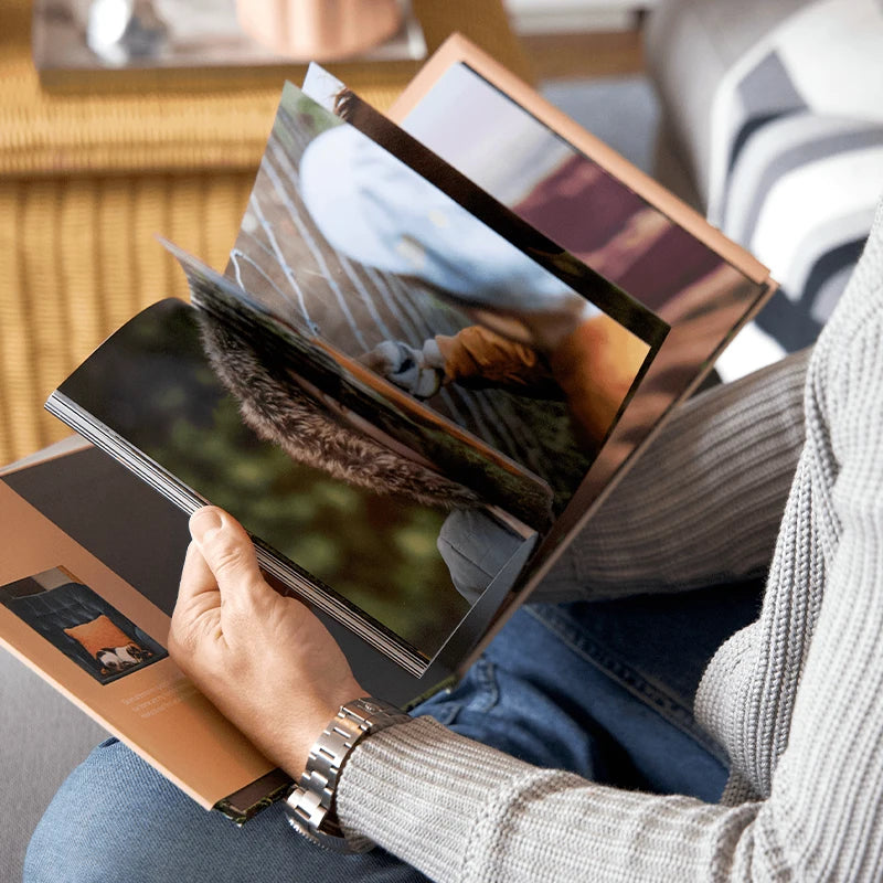Person in a gray knit sweater flipping through the pages of a hardcover photo book while seated on a sofa.