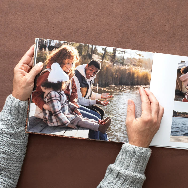 Hands holding open a hardcover photo book with a family photo of two parents and a toddler sitting on a dock by a lake in autumn.