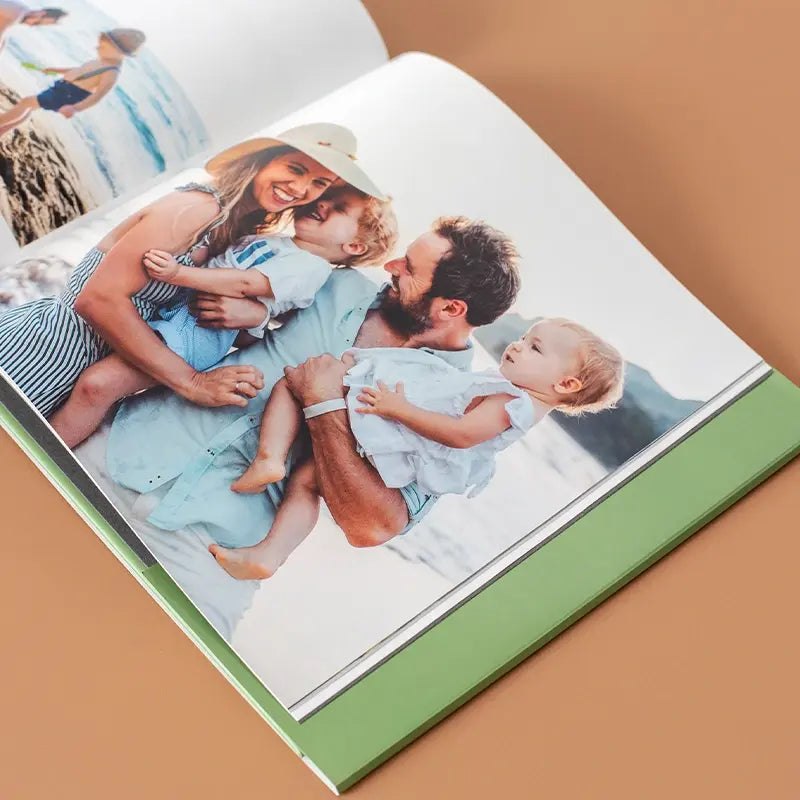 Open hardcover photo book with a green spine on a warm beige surface, displaying a full-page photo of a happy family of four laughing together on a beach