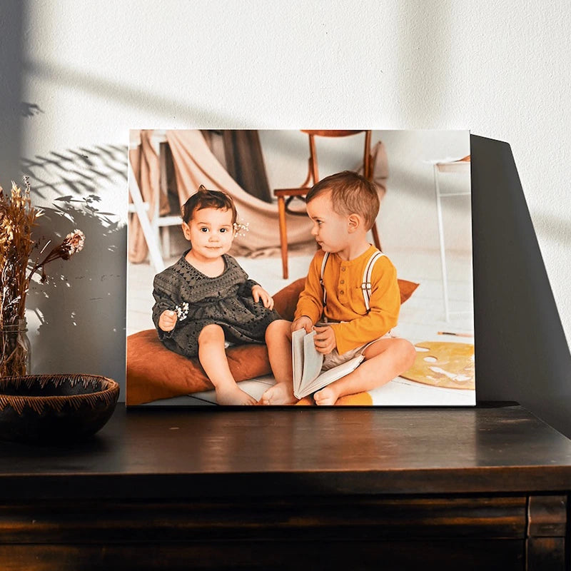 Canvas print of two young children sitting on floor cushions with a book, leaning on a wooden dresser in soft afternoon light.