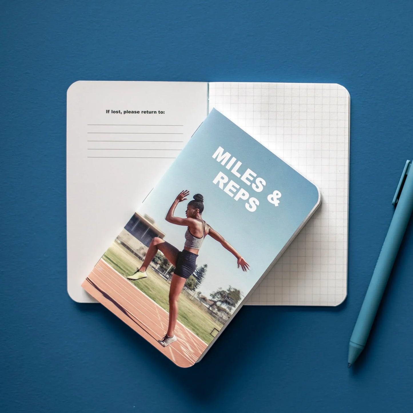 Open memo book with grid pages beneath a “Miles & Reps” fitness cover, shown on a blue background with a pen nearby.