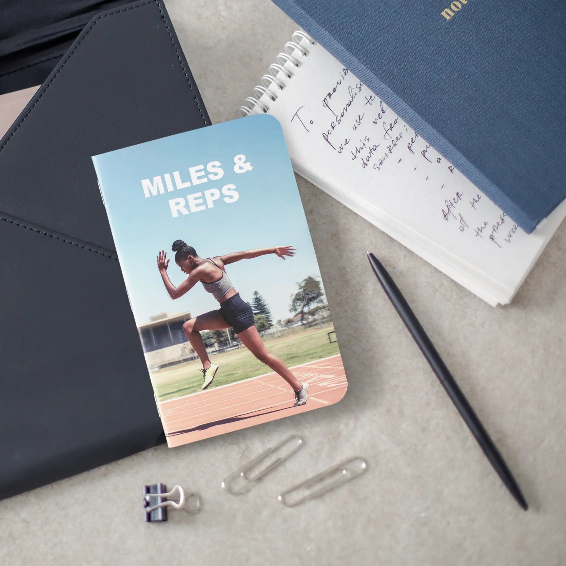Fitness-themed memo book with a runner on a track cover titled “Miles & Reps,” placed among notebooks, clips, and a pen.