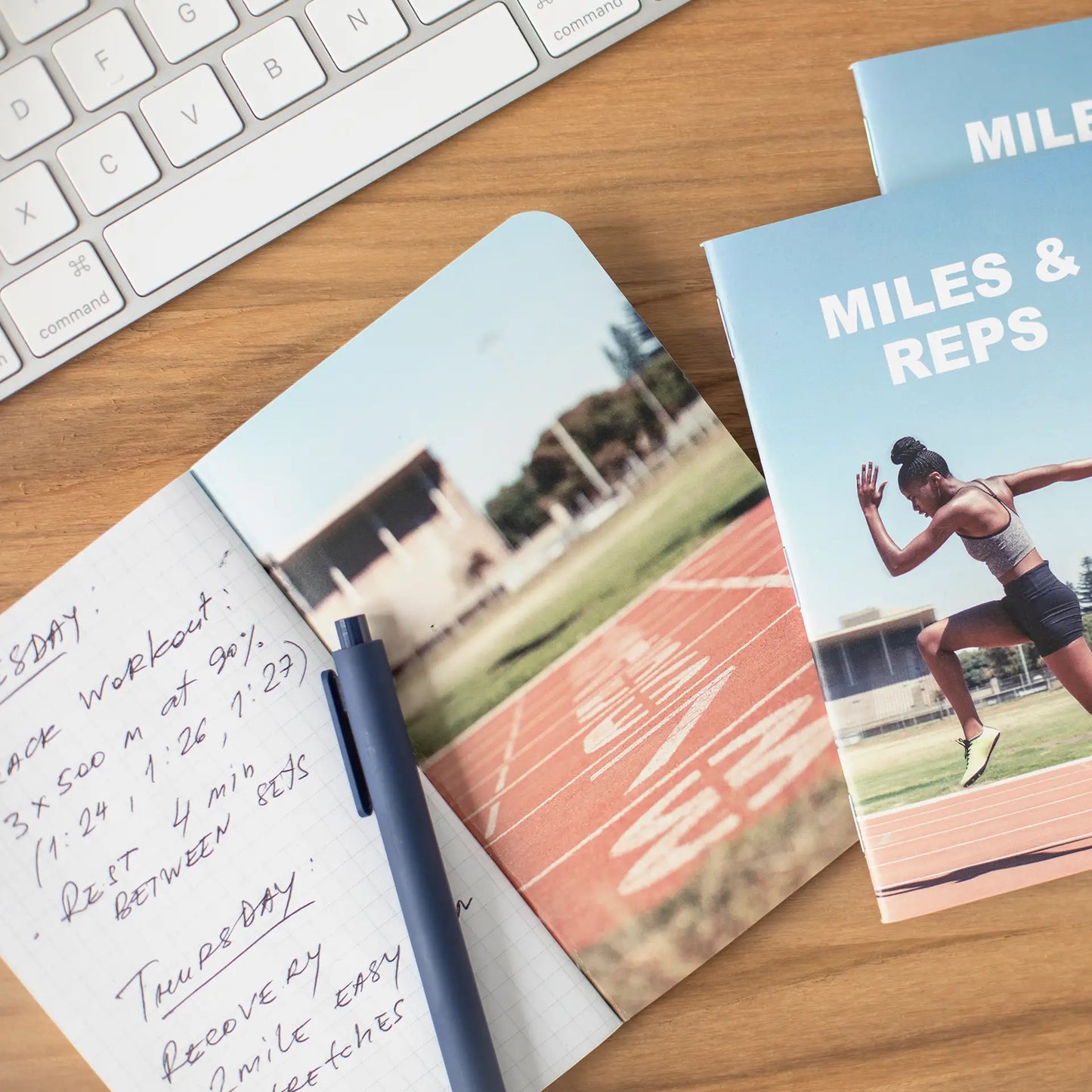 Fitness-themed memo book with a runner on a track cover titled “Miles & Reps,” placed among notebooks, clips, and a pen.