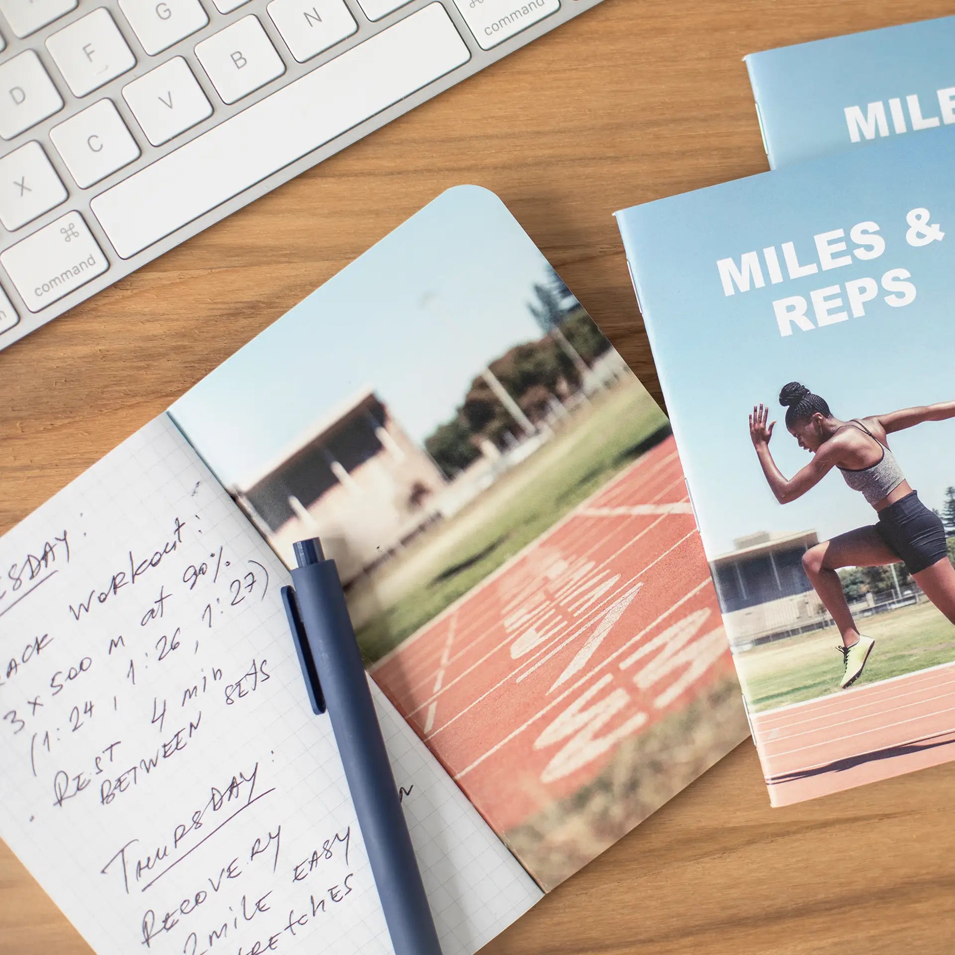 Fitness-themed memo book with a runner on a track cover titled “Miles & Reps,” placed among notebooks, clips, and a pen.