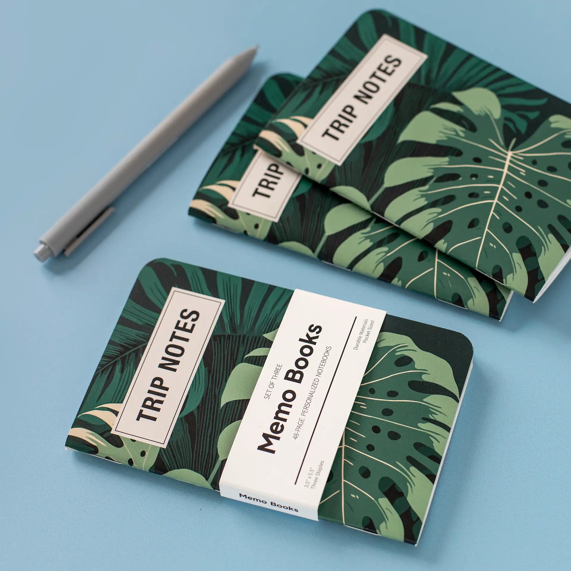Three memo books with green tropical leaf covers labeled “Trip Notes,” displayed as a stacked set on a blue background.