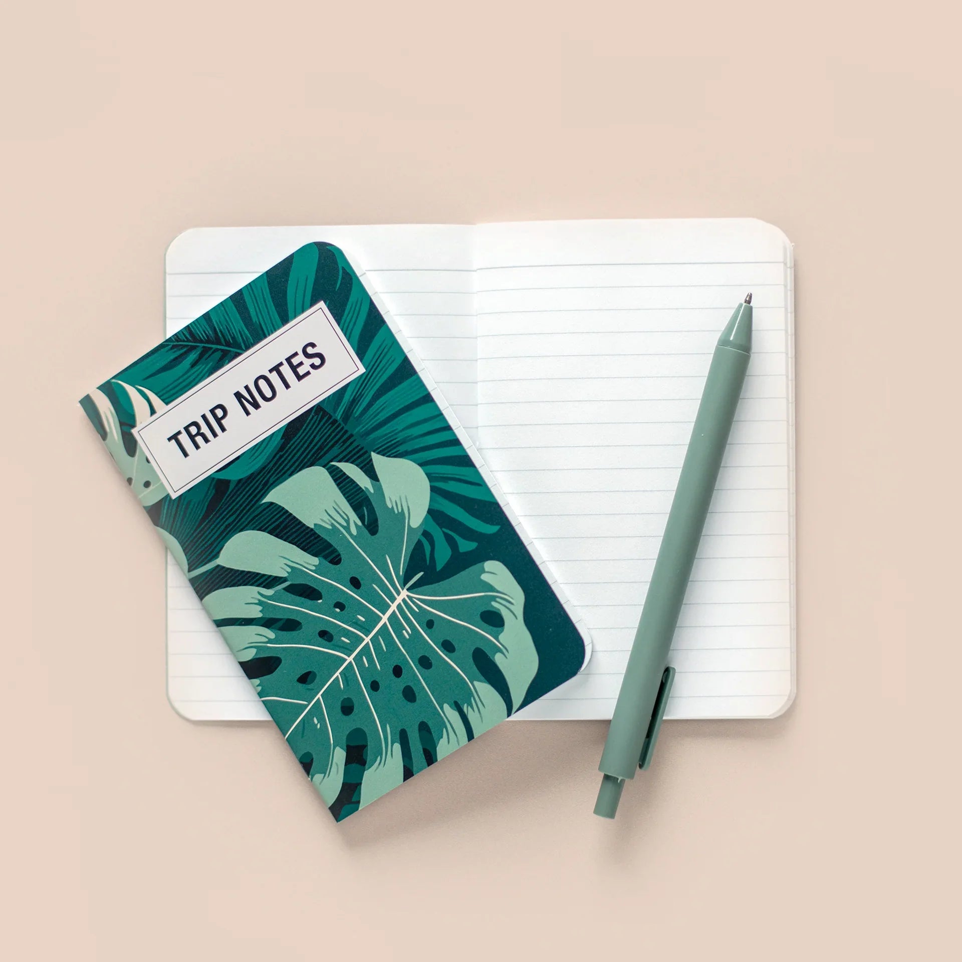 Memo book with a green tropical leaf “Trip Notes” cover resting on open lined pages, styled with a pen on a neutral surface.