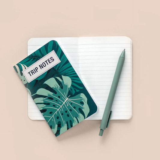 Memo book with a green tropical leaf “Trip Notes” cover resting on open lined pages, styled with a pen on a neutral surface.
