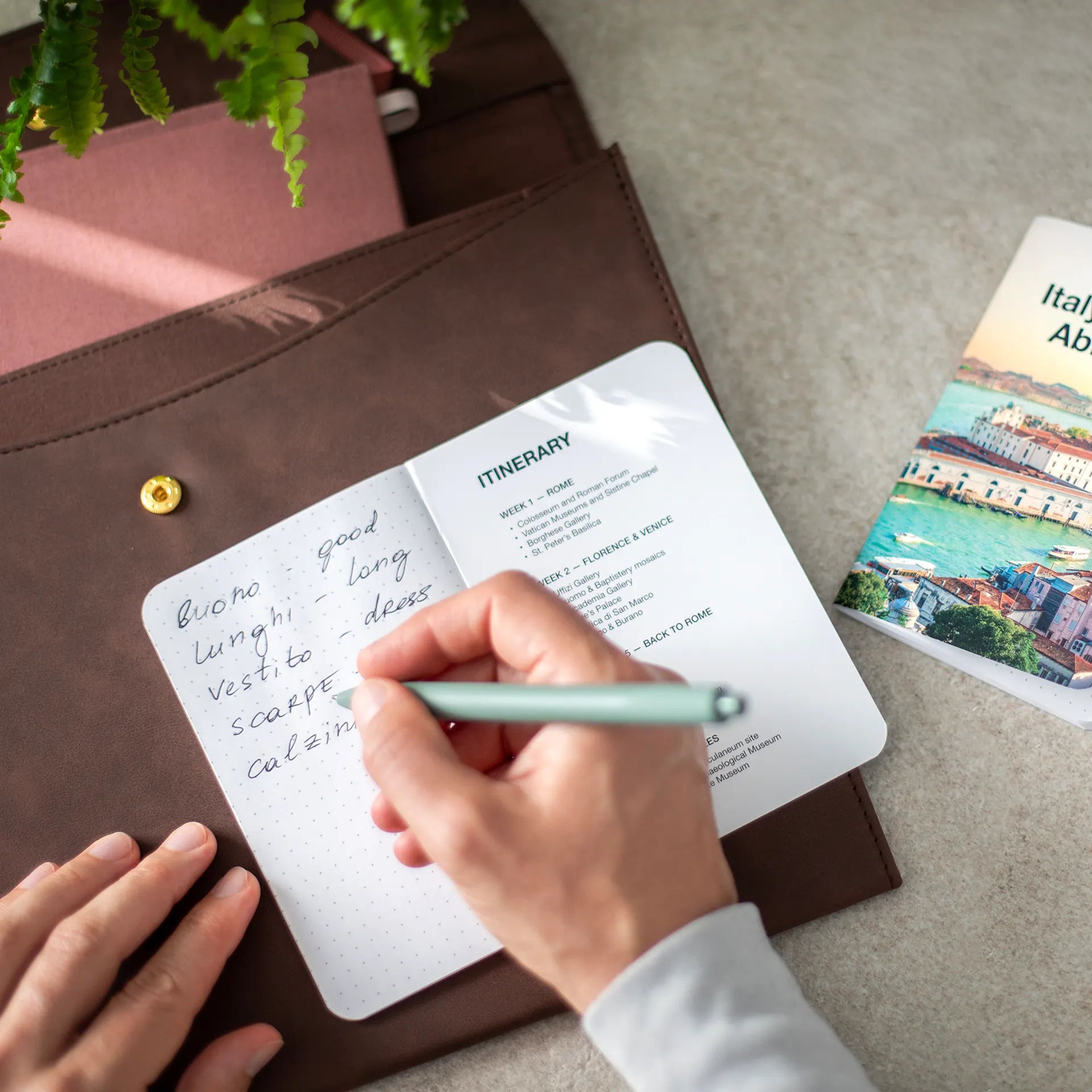 A person writing by hand in an open memo book with dotted pages, resting inside a brown leather pouch beside a travel guide.